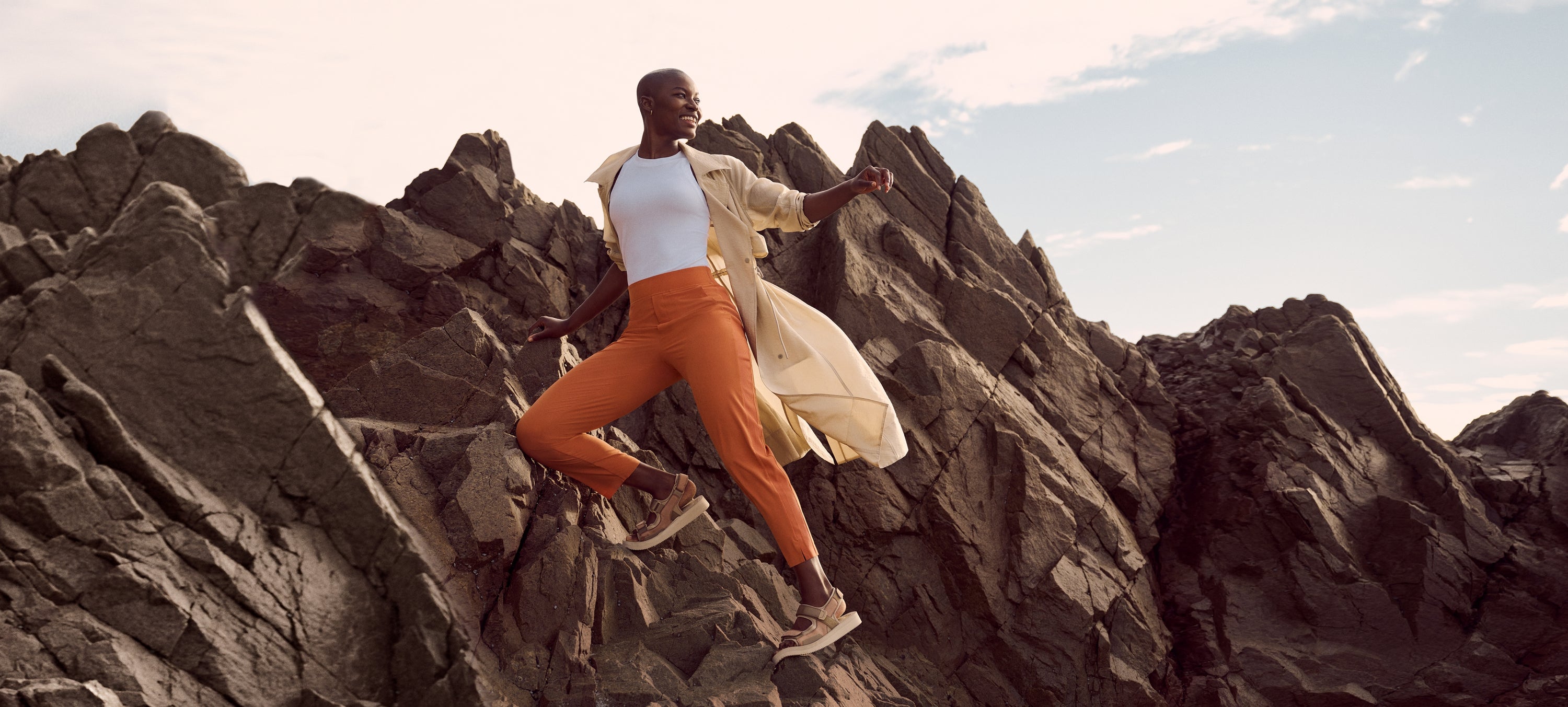 Individual climbing craggy rocks in a light yellow Athleta trenchcoat, a white Athleta tank top, orange Athleta track pants, and athletic shoes. 